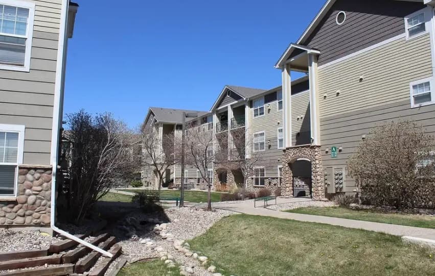 Rams Park Apartments Fort Collins feature tan and brown siding, stone accents, and balconies. Pathways wind through landscaped areas with grass, bushes, and rocks—all under a clear blue sky.