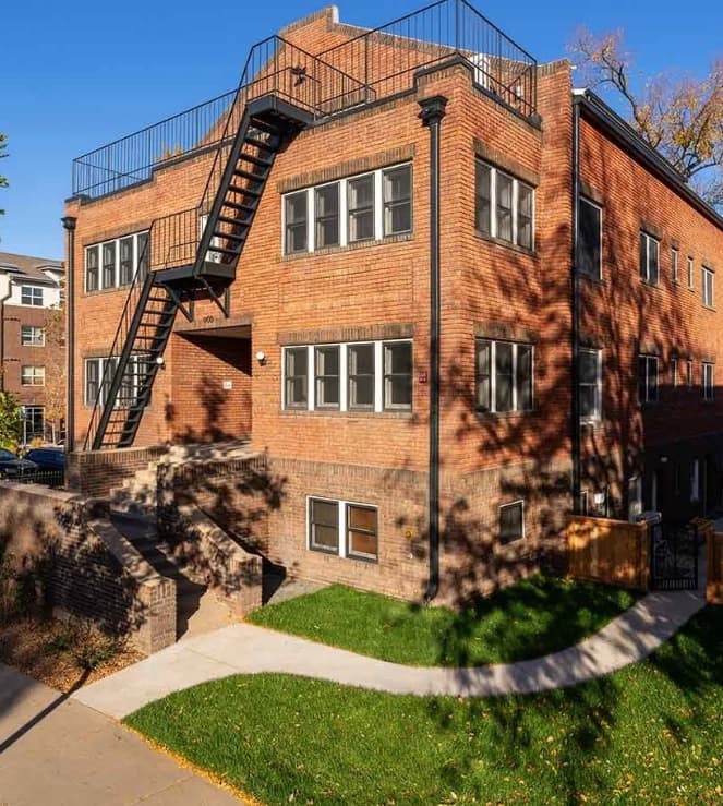 The Scott Apartments Fort Collins: A two-story red brick apartment building with large windows, an outdoor metal staircase, and a small lawn bordered by a sidewalk under a clear blue sky. Shadows of trees are visible on the building.