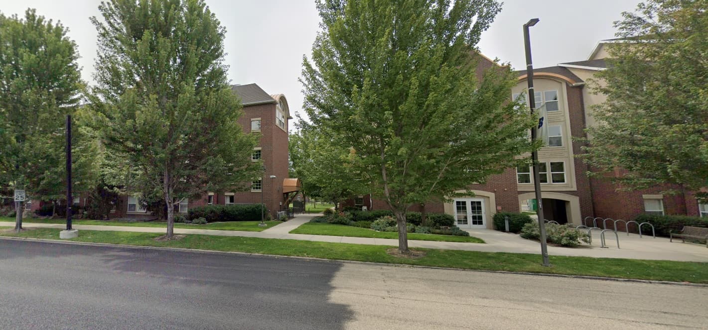 Two brick buildings, part of Selway Suites Boise, are separated by a walkway and partially hidden by tall, leafy trees. Bike racks, bushes, and a sidewalk line the sunny day with a paved road in the foreground.