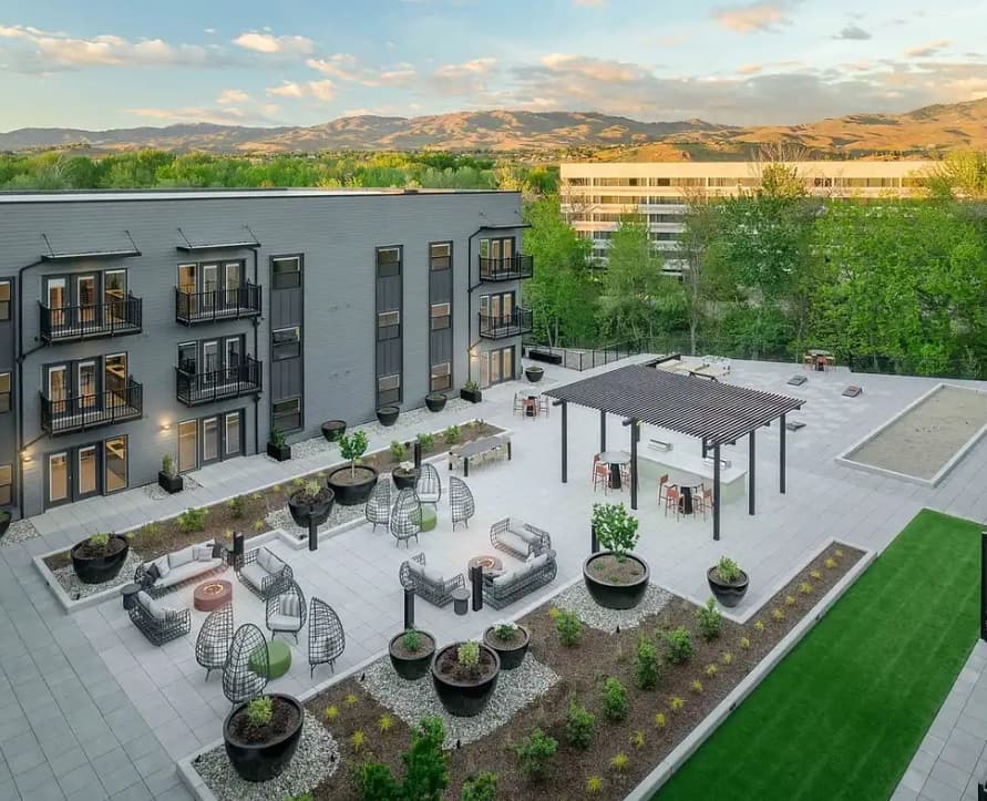 A modern rooftop patio at The Mill at Loggers Creek Apartments Boise features seating areas, fire pits, a pergola, planters, a bocce ball court, and views of green trees and mountains in the background.