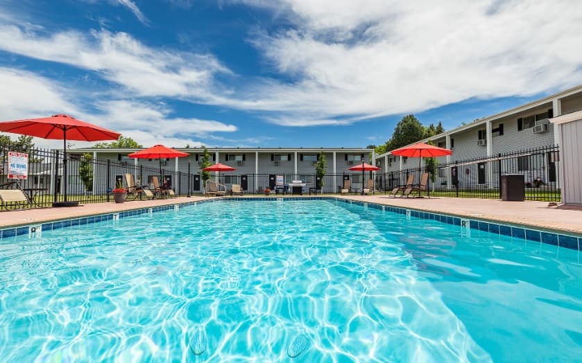 Outdoor swimming pool with clear blue water, surrounded by lounge chairs and red umbrellas at Mount Vernon Apartments Boise. Apartment buildings and a bright blue sky with scattered clouds create a relaxing backdrop.
