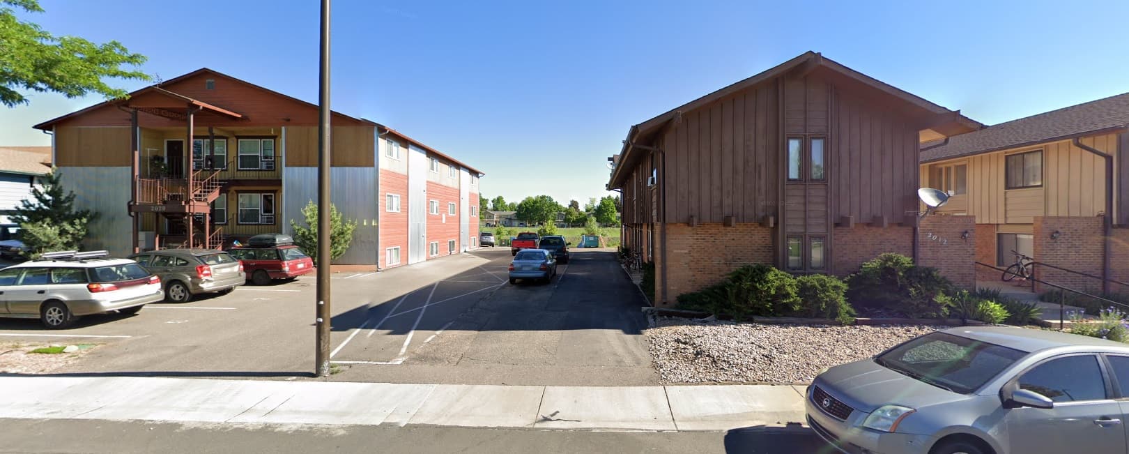Two apartment buildings with wood and brick exteriors face each other across a parking area at Village on Plum Fort Collins. A clear blue sky is visible, with trees and a grassy area in the background.