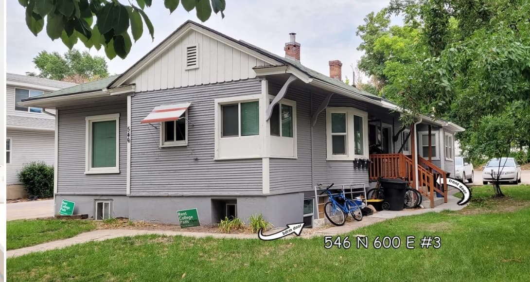 Gray single-story house with white trim and a small porch. Bicycles are next to the porch, and a white arrow points to a basement window labeled 546 N 600 E #3, part of Autumn Hills 546 Logan. Green lawn signs are visible in front.