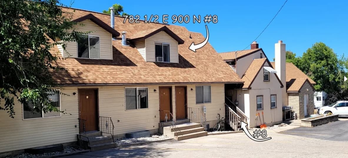 Two beige Auburn Heights townhouses with brown roofs; arrows and text label an upstairs window as 78 1/2 E 900 N #8 and a basement door as #8. Concrete steps lead to entrances, with trees, cars, and the 782 Loft Logan nearby.