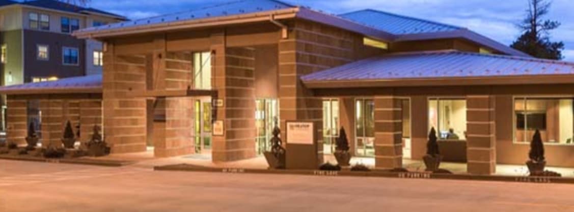 A modern, single-story building with large windows and exterior lighting at dusk sits among small trees at Hilltop Townhomes Flagstaff. A clearly marked parking lot is visible in the foreground.