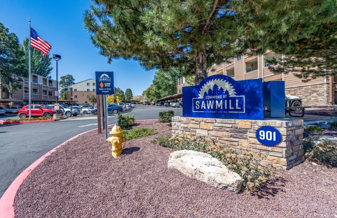 A blue sign reading Commons at Sawmill Flagstaff, 901 stands on a stone base near a fire hydrant, with a U.S. flag, trees, parked cars, and a multi-story building in the background.