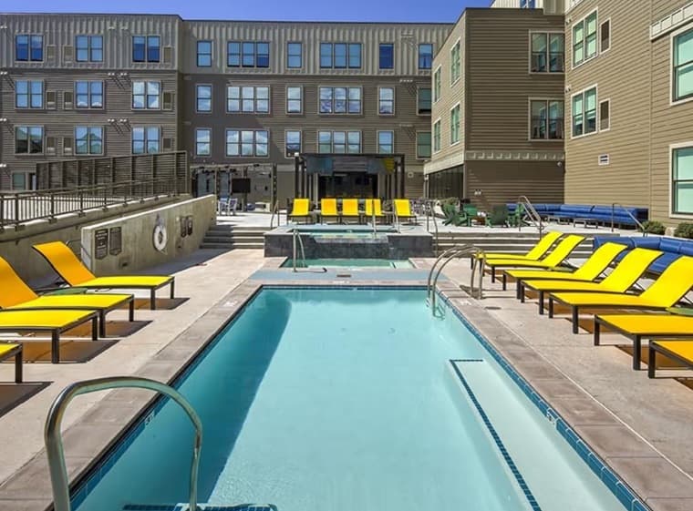 The Jack Flagstaff features an outdoor pool area with yellow and orange lounge chairs, a hot tub, and modern multi-story buildings in the background under a clear blue sky.