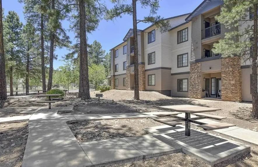 A shaded outdoor area with picnic tables, benches, and paved walkways beside The Ridge at Clear Creek Flagstaff’s three-story apartment building, surrounded by tall pine trees under a clear sky.