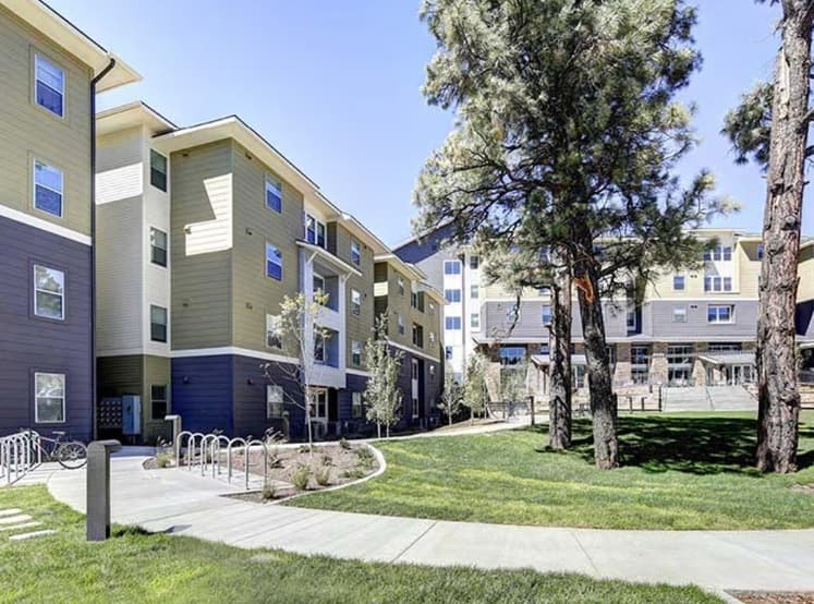 SkyView Flagstaff is a modern apartment complex with light yellow and gray siding, large windows, balconies, and surrounded by trees, green grass, and a paved walkway on a sunny day.