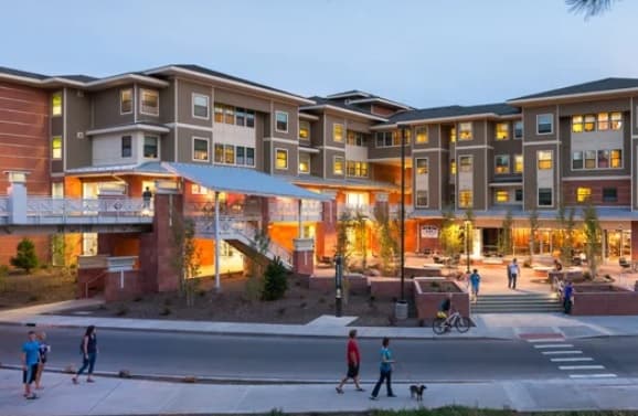 Modern multi-story student housing building, The Suites Flagstaff, glows at dusk with lit windows. People walk, bike, and stroll dogs along the street and sidewalk. Landscaped areas and a pedestrian bridge add to the vibrant scene.