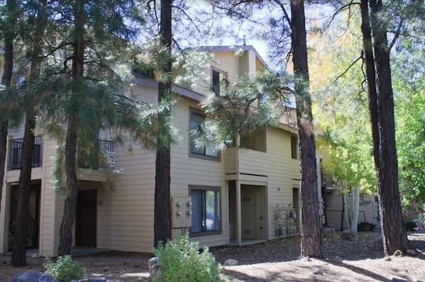 A beige, two-story apartment building with balconies in University West Flagstaff, surrounded by tall pine trees and leafy greenery on a sunny day.