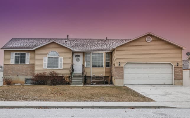 A single-story tan house at 919 W 1150 S features a white garage door, white trim, small front steps, and a brown shingle roof. It sits on a lawn with a driveway beneath a sky that fades from purple to pink.