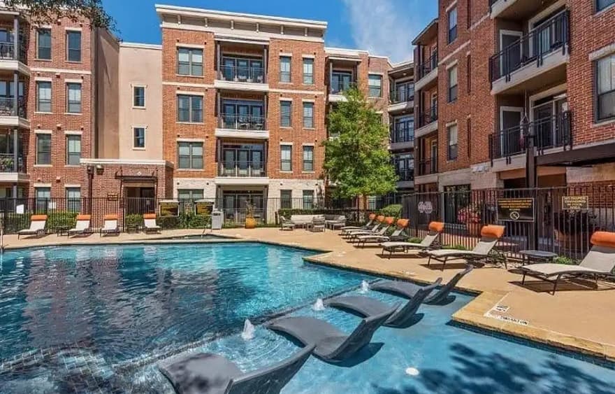 Modern Trinity Urban Apartments - Bluff & District Fort Worth complex with red brick exterior, balconies, and large windows surrounds an outdoor pool area featuring built-in lounge chairs, water jets, umbrellas, and plenty of poolside seating.