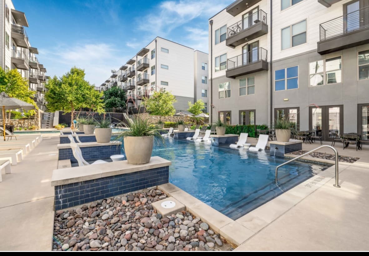 Modern apartment complex courtyard at The Bowery at Southside Fort Worth, featuring a swimming pool with partly submerged lounge chairs, potted plants, and surrounding multi-story buildings under a bright blue sky.