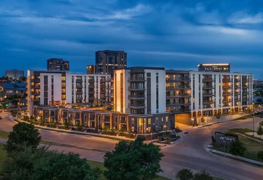 Modern multi-story apartment complex, The Braden on Fifth Fort Worth, features illuminated windows at dusk, surrounded by trees and roads, with taller buildings in the background beneath a cloudy blue sky.