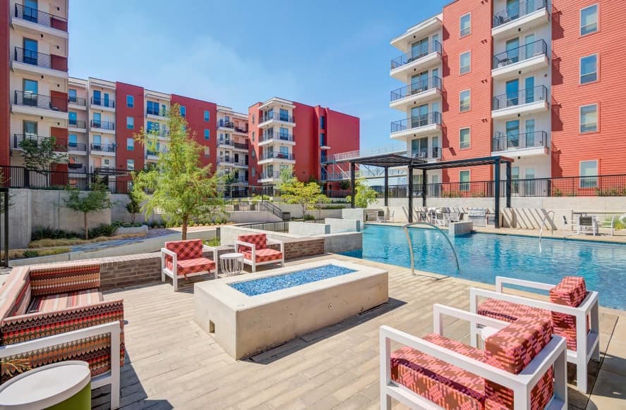 Modern apartment complex at Encore Panther Island Fort Worth, featuring striking red buildings, a swimming pool, poolside seating with red patterned chairs, a concrete fire pit, and lush landscaping under a clear blue sky.
