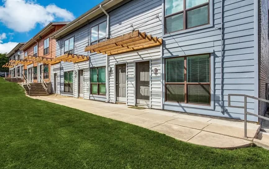 A row of modern townhomes at The Foundry Fort Worth features light blue and pink siding, large windows, wooden awnings, and a well-maintained green lawn along a concrete walkway under a partly cloudy sky.