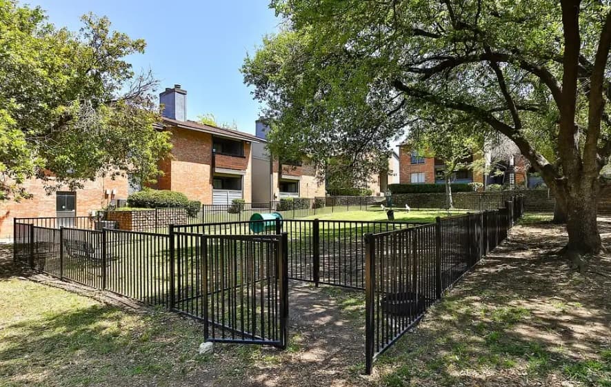 A fenced dog park with a gate, grass, and shade trees awaits outside The Haven at Chisholm Trail Fort Worth's brick apartment buildings under a clear blue sky.