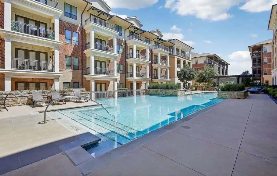 Modern apartment complex, The Kelton at Clearfork Fort Worth, features balconies overlooking a clear swimming pool, with lounge chairs and lush landscaping under a bright, partly cloudy sky.
