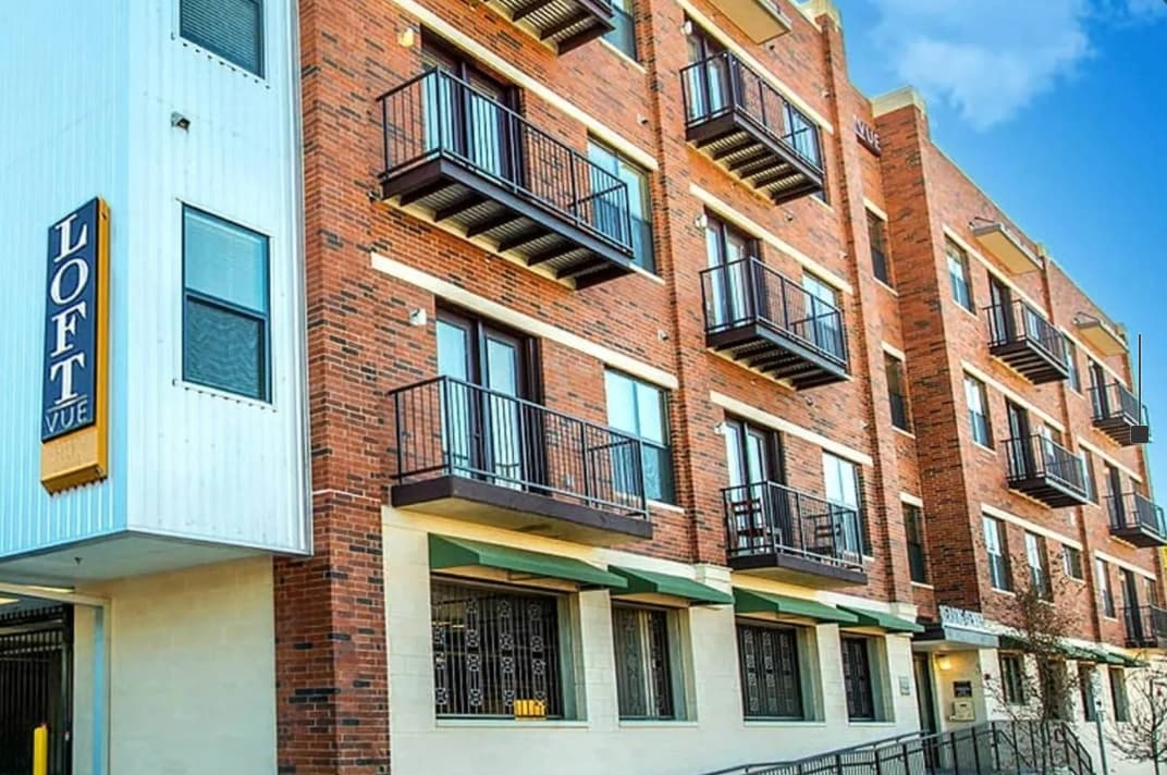 A modern brick apartment building with multiple balconies, large windows, and a sign reading LOFT VUE on the exterior. Experience Loft Vue Living Fort Worth, where green awnings grace the lower level and blue skies stretch above.
