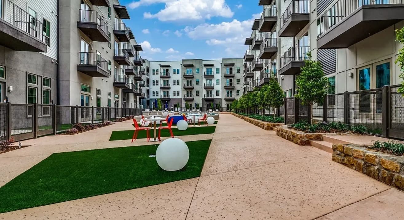 A modern courtyard at Luxia River East Apartments Fort Worth features artificial grass, round white lights, red and white chairs and tables, and multi-level balconies under a partly cloudy sky.