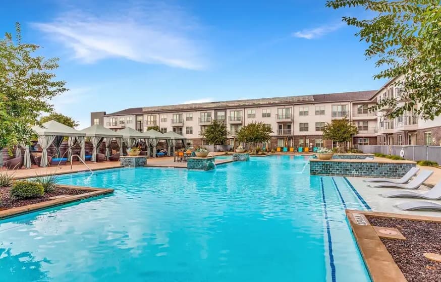 Relax at the large outdoor swimming pool at Madera at LeftBank Fort Worth, featuring lounge chairs, shaded cabanas, lush potted plants, and a multi-story apartment building under a clear blue sky.