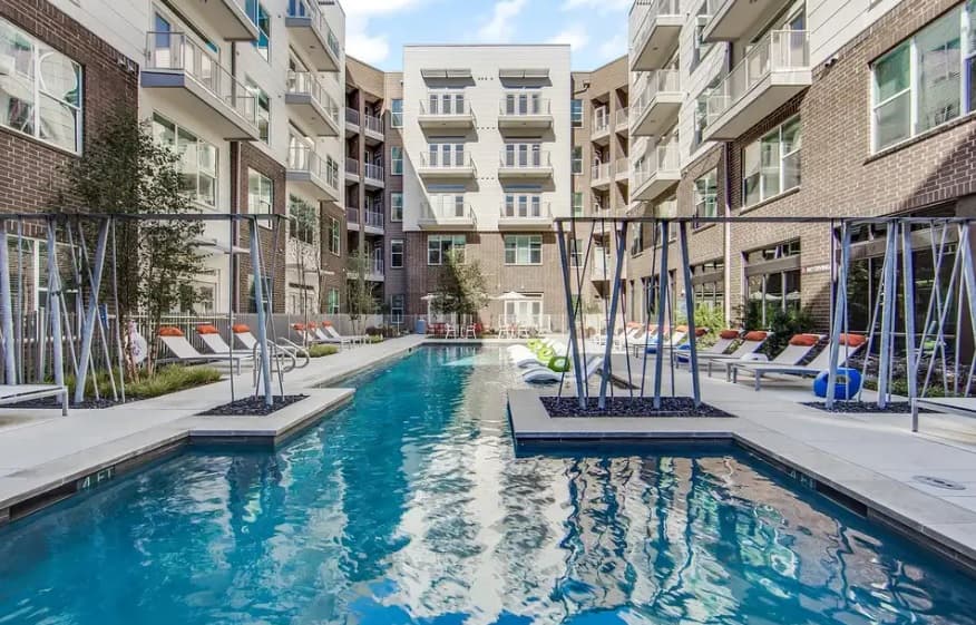 Modern apartment complex courtyard at The Monarch Medical District Apartments Fort Worth, featuring a long rectangular swimming pool surrounded by lounge chairs and lush greenery. Several balconies overlook the pool, with buildings rising four to five stories tall.