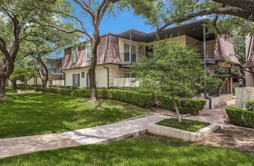 Monticello Apartments Fort Worth offers a charming two-story building with brown mansard-style roofs, beige walls, balconies, and staircases amid lush green lawns, bushes, trees, and a concrete walkway under a clear blue sky.