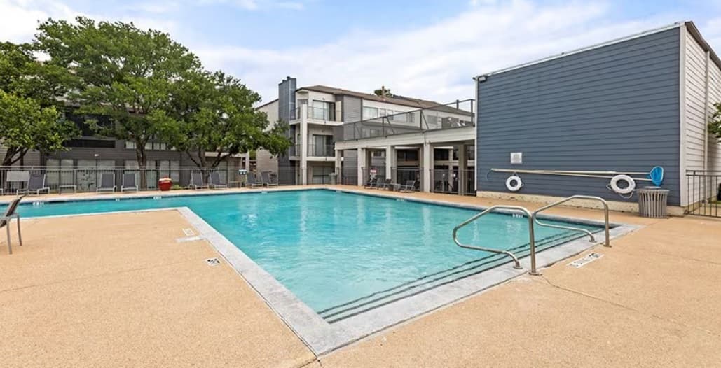 A rectangular outdoor swimming pool with metal handrails sits on a beige concrete deck at Mylo Apartment Homes Fort Worth. Apartment buildings and green trees are in the background under a partly cloudy sky.