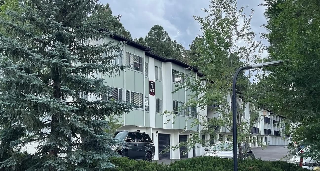 A row of three-story modern townhouses, known as Skyline Lofts Flagstaff, with white and green exteriors stands among trees and parked cars beneath a cloudy sky. A black street lamp is visible in the foreground.