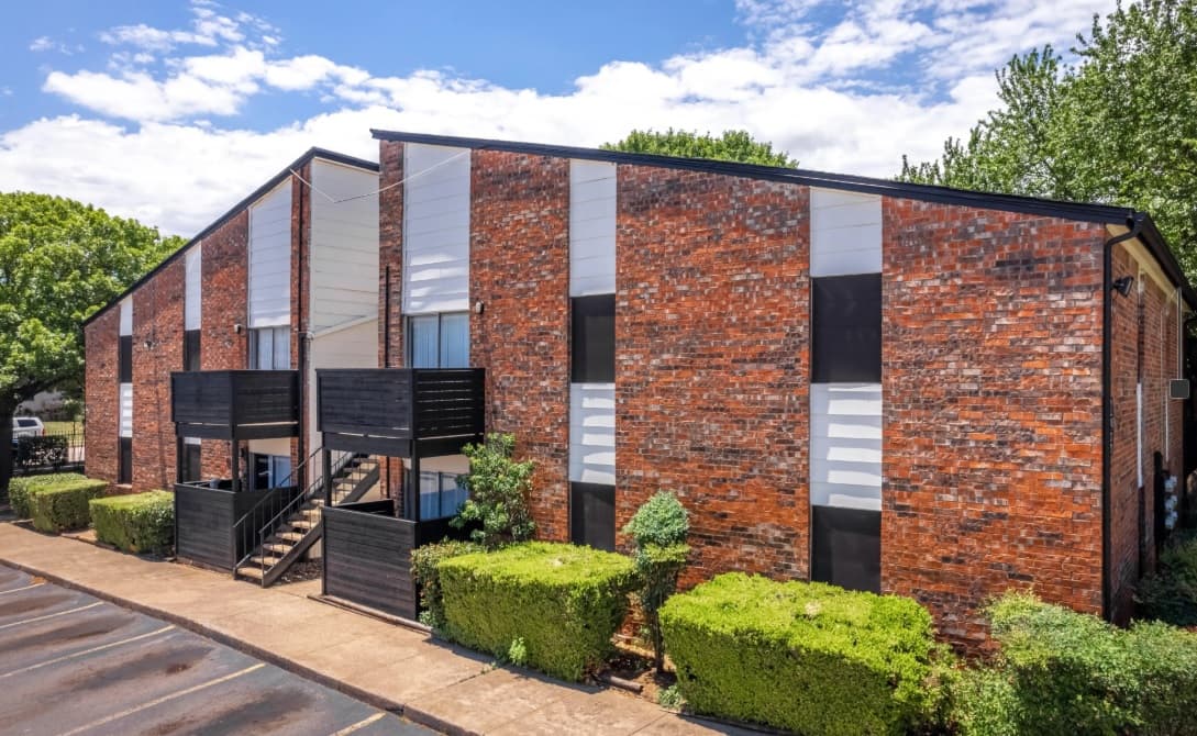 A two-story brick apartment building at Slate at Fort Worth Apartments, with black and white accents, outdoor staircases, black railings, trimmed green bushes, and trees under a partly cloudy blue sky in Fort Worth.