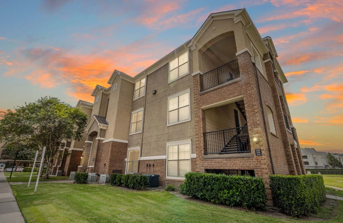 Three-story apartment building with brick and beige siding, exterior staircases, and covered balconies at The Club at Stonegate Apartments Fort Worth. Manicured lawn and bushes surround the building under a colorful sunset sky.
