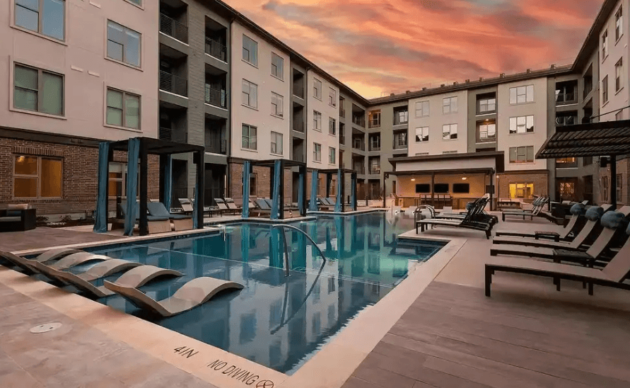 Modern apartment complex courtyard at The Elm at River Park Fort Worth featuring a rectangular swimming pool, poolside lounge chairs, shaded cabanas, and a sunset sky. Surrounding buildings rise four stories tall, creating a stylish urban retreat.