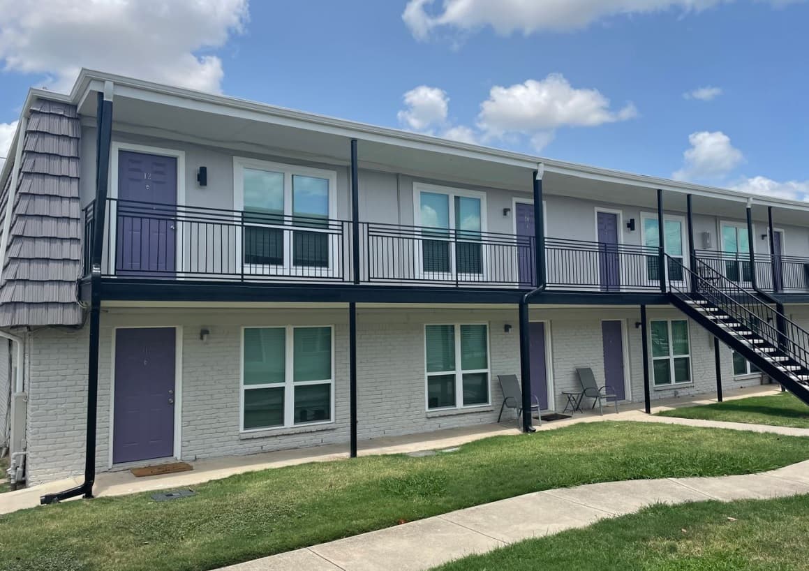University Villas Apartments Fort Worth features a two-story building with white brick walls and purple doors, an outdoor staircase, sleek black railings, and a small grass lawn in front under a partly cloudy sky.