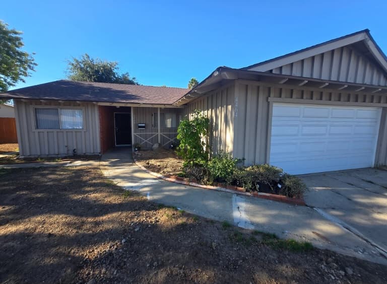 Charming single-story house on 4464 Arendo Dr with beige siding, a white double garage door, and a small front porch. Plants and shrubs frame the entrance, while the yard features bare soil patches and a curved concrete walkway.