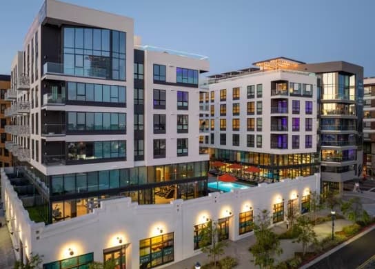 Modern multi-story apartment buildings on Gala Ave with large windows surround a central courtyard featuring a swimming pool. The scene is taken at dusk, with lights illuminating the buildings and lush landscaping below.