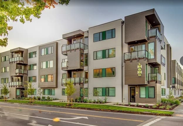 The Andy Eugene is a modern three-story apartment building with a gray and white exterior, large windows, balconies, and green landscaping along a quiet street.