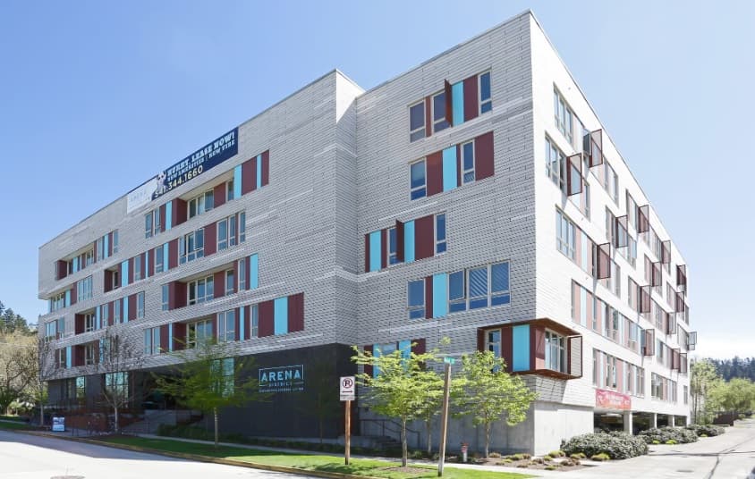 A modern multi-story building, Arena District Apartments features a white brick exterior, red window accents, large glass windows, and street trees in front, all captured on a sunny day.