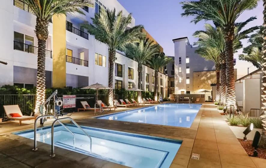 A modern outdoor swimming pool area at Array La Mesa, lined with palm trees, lounge chairs, and umbrellas, surrounded by multi-story buildings at dusk with soft lighting.