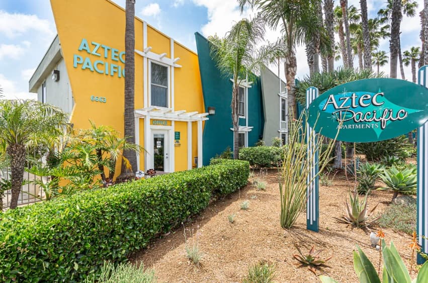 Aztec Pacific Apartments features a bright yellow and teal exterior, surrounded by palm trees and landscaped plants, with a large green sign showcasing the Aztec Pacific Apartments name out front.