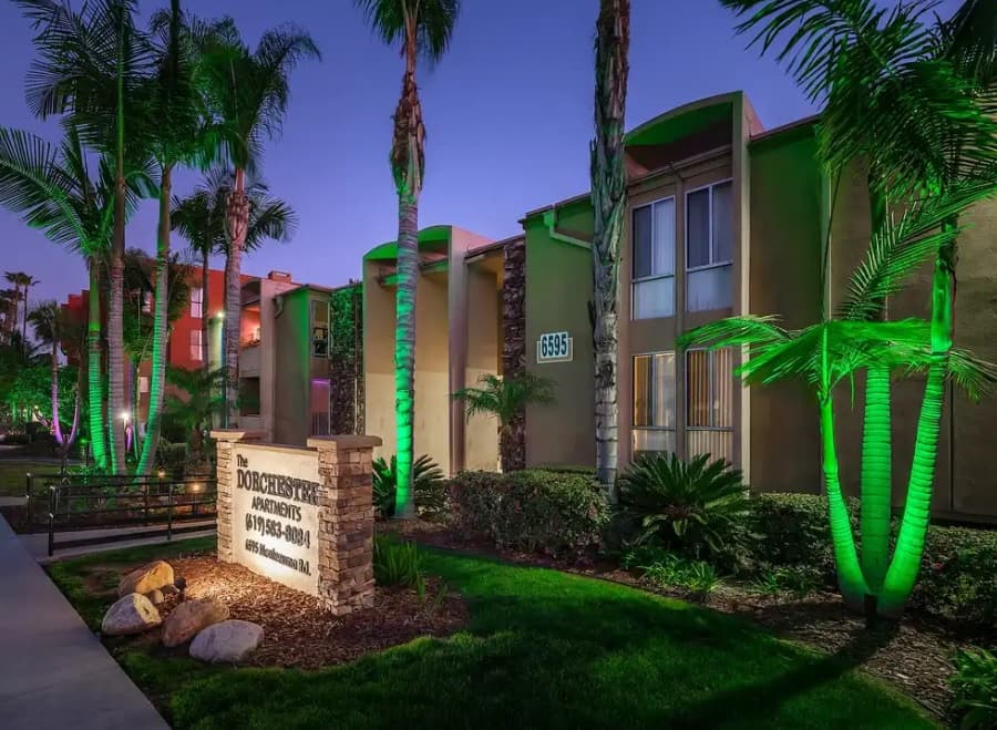A modern apartment building with tall windows and palm trees, illuminated by green and purple lights at dusk. A lit sign in front reads The Dorchester Apartments with a phone number.