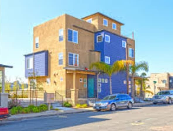 A modern three-story townhome apartment building in Fresno with tan and blue exterior walls, large windows, palm trees, and parked cars lining the street on a sunny day.
