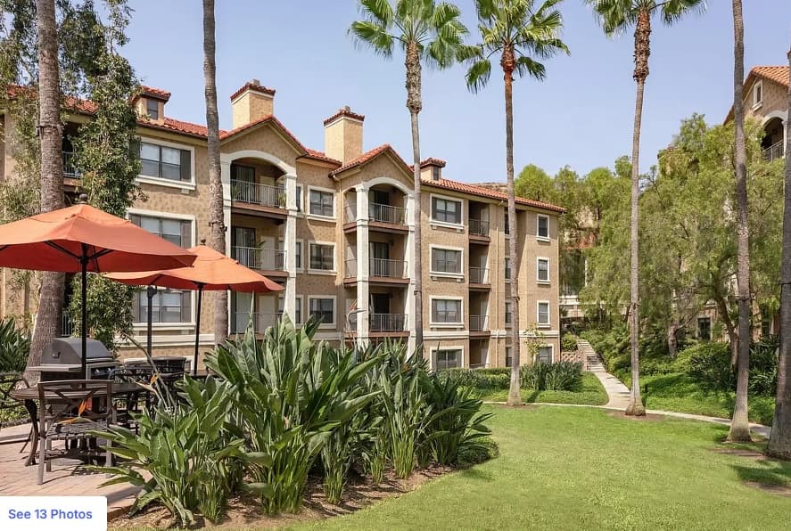 A multi-story apartment building with balconies and red tile roofs is surrounded by tall palm trees, green lawns, and lush plants. There is a shaded outdoor patio area with tables and orange umbrellas in the foreground.