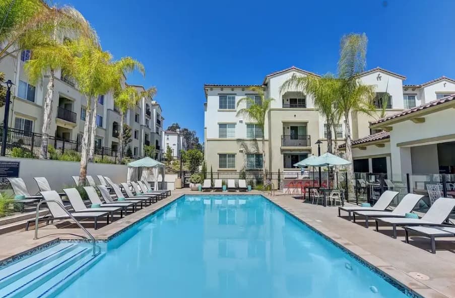 A modern apartment complex with palm trees, a swimming pool, lounge chairs, and umbrellas under a clear blue sky. The buildings are three stories with balconies and light-colored exteriors.