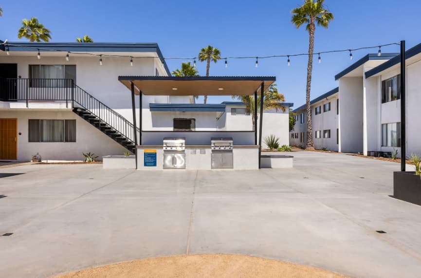 Outdoor communal area in an apartment complex featuring two stainless steel grills under a shaded pergola, surrounded by white two-story buildings, palm trees, and string lights overhead.