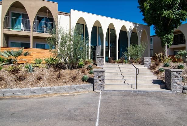 A modern building with tall, arched columns at the entrance, Veranda La Mesa features drought-tolerant landscaping and stone planters. Concrete steps with a handrail lead up to glass doors, while balconies are visible on the left.