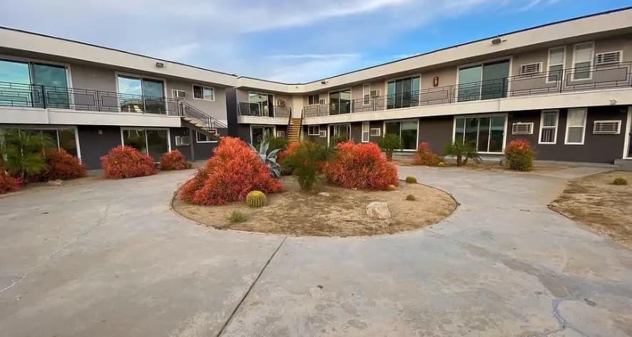 Willow Glen Apartments features a two-story complex encircling a circular courtyard with red bushes, cacti, and dry soil. Concrete walkways lead to glass sliding doors and metal railings beneath a partly cloudy sky.