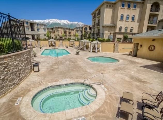 Outdoor view of an apartment complex reminiscent of Italian Villages, featuring a hot tub in the foreground, two swimming pools, lounge chairs, cabanas, and mountain scenery in the background under a clear blue sky.