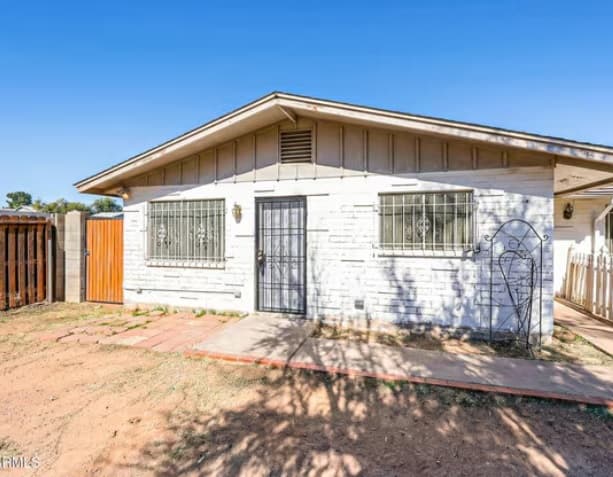 A single-story white brick house at 2201 W Heatherbrae Dr, Phoenix, AZ 85015, featuring barred windows, a metal security door, wooden gate, small garden area with sparse grass, and a clear blue sky in the background.