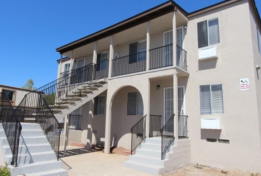 A two-story beige apartment building on Jessie Avenue in La Mesa, featuring two exterior staircases with black railings, multiple windows with blinds, and a sign on the wall, all under a clear blue sky.